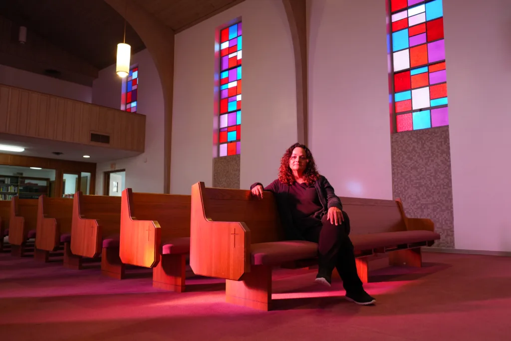 A woman sits in a pew of a church. Stained glass windows illuminate her face.